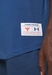 Blauwe sportshirt heeft een wit label met rode accenten, waarop een stierlogo en de merknaam "UNDER ARMOUR" zijn gestikt op de zoom.