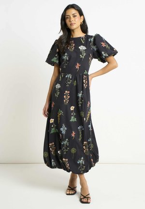 Woman wearing a black floral midi dress with puff sleeves and balloon hem, paired with black strappy open-toe heels, standing against a white wall.