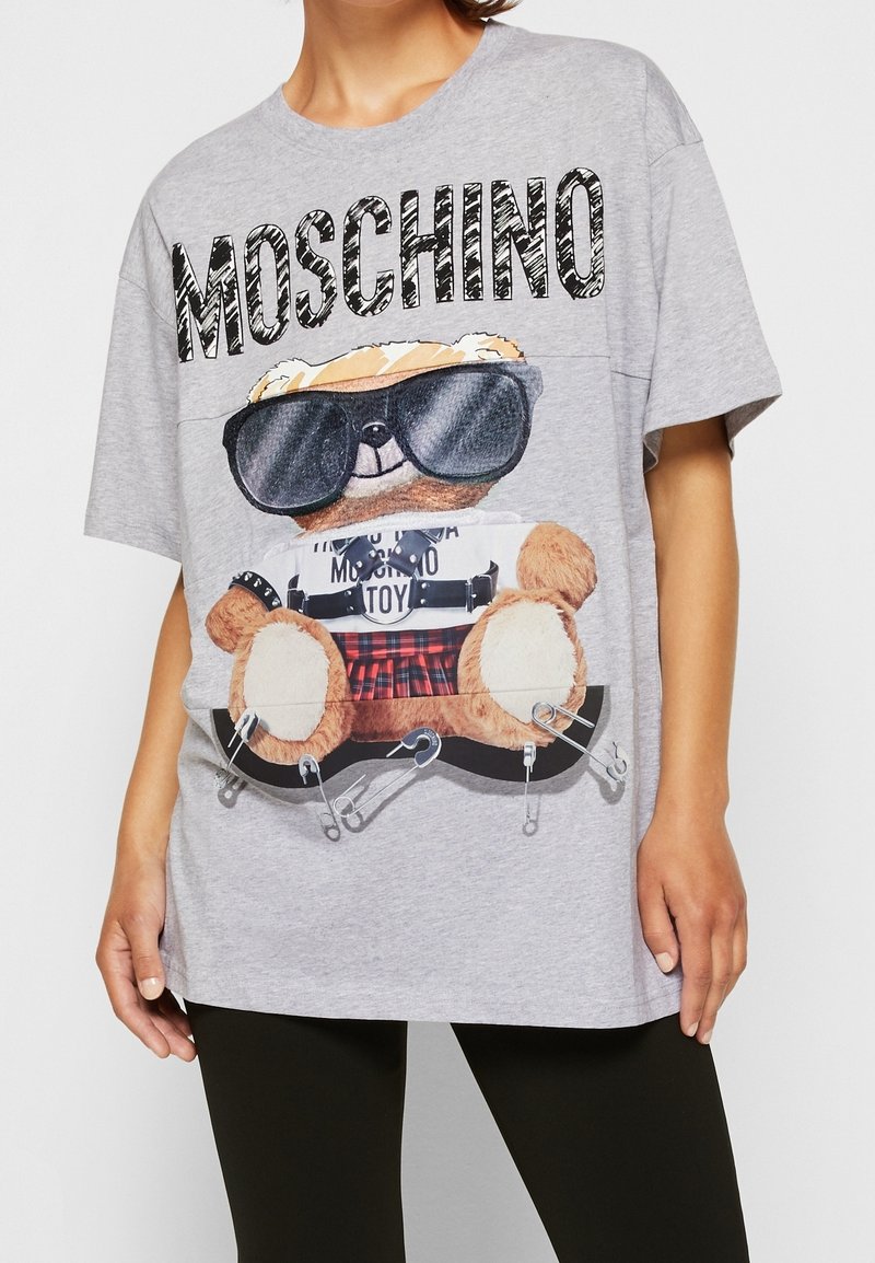 Grå oversized t-shirt med en stor grafik av en björn i solglasögon, med säkerhetsnålar och design av rutigt kjol. Varumärket "Moschino" framträdande visat.