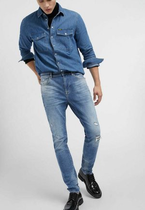 Mann trägt ein blaues Jeanshemd mit hochgekrempelten Ärmeln, hellblaue zerrissene Jeans und schwarze Schnürschuhe, steht vor einem schlichten Hintergrund.