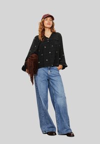 Femme portant un blouse florale noire, un jean bleu à jambes larges, une casquette bordeaux, tenant un sac en cuir frangé marron, se tenant avec confiance contre un fond gris.