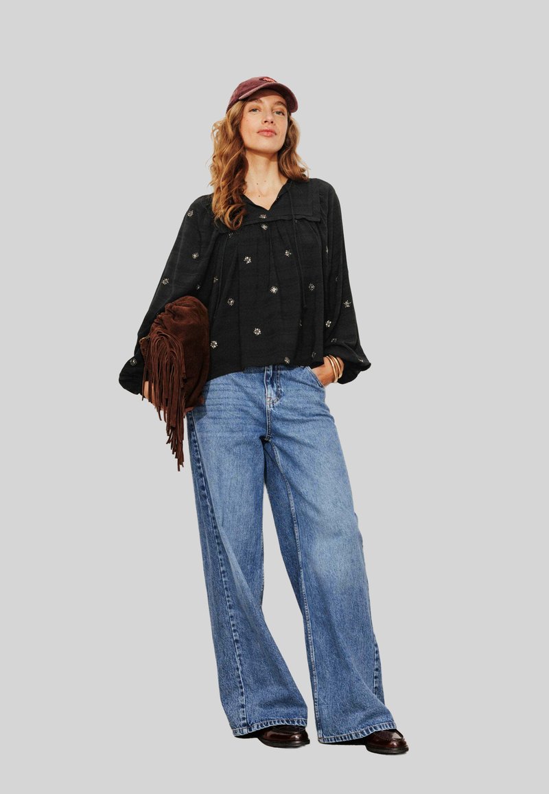 Femme portant un blouse florale noire, un jean bleu à jambes larges, une casquette bordeaux, tenant un sac en cuir frangé marron, se tenant avec confiance contre un fond gris.