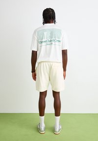 T-shirt blanche avec un graphisme vert, "Ellesse Club di Corsa," associée à un short beige clair. Le modèle porte des chaussures argentées et des chaussettes blanches.
