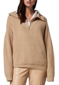 Pull en tricot beige à côtes avec un col zippé demi, épaules tombantes, associé à un pantalon ajusté de couleur claire. Détails de tissu texturé visibles.