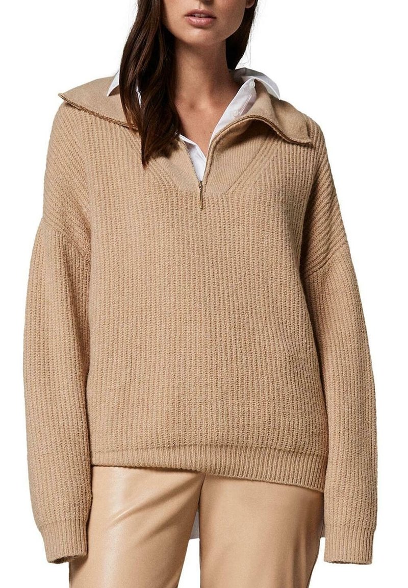 Pull en tricot beige à côtes avec un col zippé demi, épaules tombantes, associé à un pantalon ajusté de couleur claire. Détails de tissu texturé visibles.