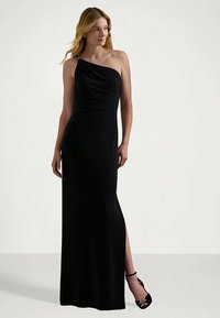 Ženska v dolgi črni obleki z enim ramenom in stranskim razporkom, ki nosi črne sandale s peto, stoji ob enobarvni podlagi.