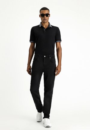 Schwarzes Polohemd mit grauen Kragenakzenten, kombiniert mit dunklen Jeans und weißen Sneakers. Das Modell trägt Sonnenbrillen. Gerade geschnittenes Design.