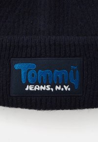Mornarska pletena kapa s pravokutnom etiketom na kojoj piše "Tommy Jeans, N.Y." u plavoj i bijeloj vezu. Mekana tekstura, rebrasti rub.