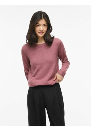 Ung kvinde med skulderlangt mørkt hår iført en støvet pink sweater og sorte viddebenede bukser, stående mod en ensfarvet hvid baggrund.