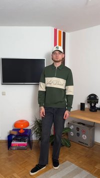 Zelena zip-up jopica s kremasto črto, ki ima izvezen napis, v kombinaciji s temno modrimi kavbojkami in črnimi natikači. Preprost, funkcionalen dizajn.