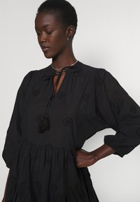 Black dress with a tie neck, featuring embroidery and tassels. Long puff sleeves and tiered design, made of lightweight fabric.