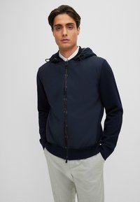 Marinblå zip-up hoodie med en mjuk tyghuva, kontrasterande ärmar och elastiska cuffar. Har dragsko och en slät dragkedja.