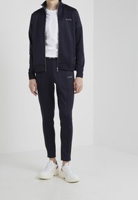 Námorníky modrý sportovní set s bundou a kalhotami v hladké látce, s upevněním na zip, žebrovanými manžetami a bílým názvem značky "LES DEUX". V kombinaci s bílými teniskami.