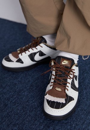 Baskets Nike blanches, noires et marron avec des rayures fines et des franges décoratives en cuir, portées avec un pantalon beige et des chaussettes blanches sur un tapis bleu.