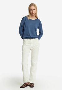 Blå långärmad topp i kombination med vita rakbeniga byxor och bruna sandaler med remmar; har en rundad halsringning och avslappnad passform.