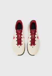 Nike løbesko med en cremefarvet mesh-overdel, røde snørebånd og detaljer, sort indmad og en struktureret sort gummisål. Bemærkelsesværdig logo-detalje på siden.