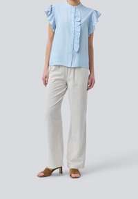Mujer vistiendo blusa sin mangas de volantes en un tono azul claro, pantalones blancos a rayas verticales y sandalias de tacón abierto en color marrón, de pie sobre un fondo gris.