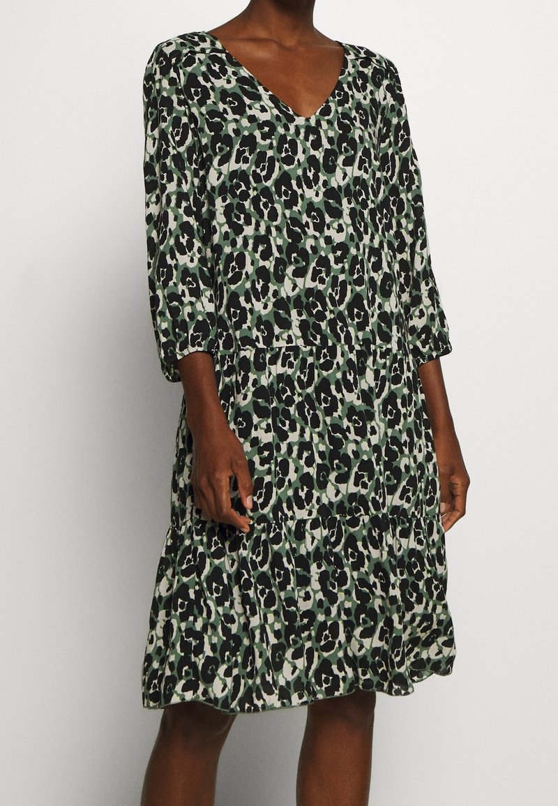 Femme portant une robe longueur genou avec un imprimé léopard abstrait noir, vert et beige, et des manches trois-quarts sur un fond uni.