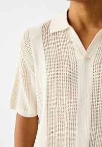 Polo de color crema con un diseño texturizado y acanalado que presenta patrones verticales de tejido abierto. Tela suave, mangas cortas y un cuello en forma de V.