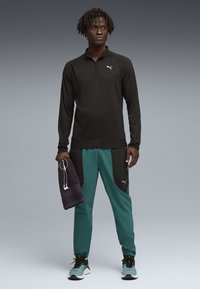 Άνδρας που φοράει μαύρο μπουφάν με φερμουάρ, γαλάζιες και μαύρες φόρμες, μπλε αθλητικά παπούτσια, κρατώντας μια σκοτεινή πετσέτα με άσπρα ακουστικά που κρέμονται από αυτήν.