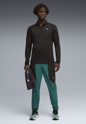 Vyras juodu užtrauktuku užsegamu megztiniu, žalsvai mėlynomis ir juodomis sportinėmis kelnėmis, mėlynais sportbačiais, laiko tamsią rankšluostį, ant kurio kabo balti ausinukai.