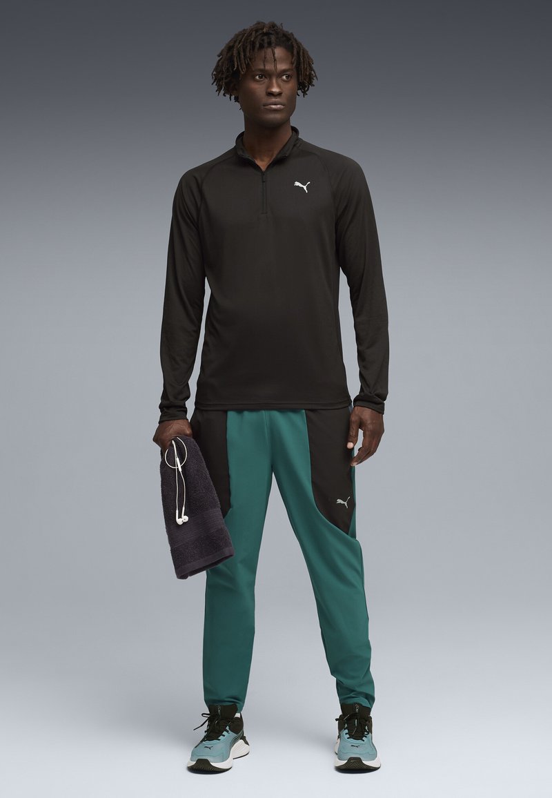 Άνδρας που φοράει μαύρο μπουφάν με φερμουάρ, γαλάζιες και μαύρες φόρμες, μπλε αθλητικά παπούτσια, κρατώντας μια σκοτεινή πετσέτα με άσπρα ακουστικά που κρέμονται από αυτήν.