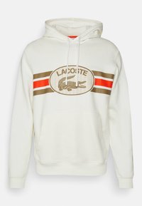 Felpa bianca con tasca a marsupio, caratterizzata da strisce orizzontali marroni e rosse e un grande logo Lacoste con emblematico coccodrillo.