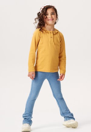 Senf-farbenes Langarmshirt mit Knopfdetails und Rüschenausschnitt, kombiniert mit hellblauen Jeans aus elastischem Material und weißen Sneakers.