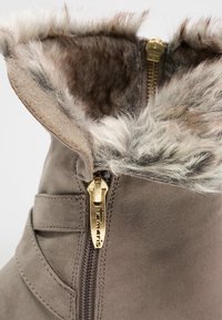 Brun ruskind støvle med kunstpelsforing foroven, guld lynlåsdetalje og en struktureret overflade. Accenter inkluderer et dekorativt syningsdesign.