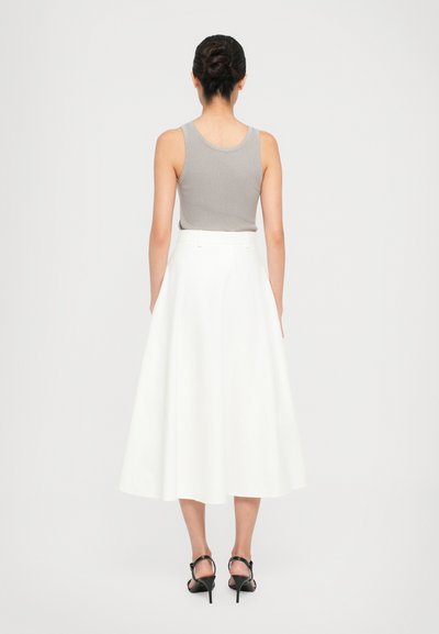 Woman with hair in a bun wearing a gray sleeveless top, white midi skirt, and black high-heeled sandals standing against a plain background.