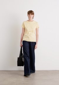 T-shirt di cotone gialla con scollatura rotonda, abbinata a jeans in denim a gamba larga blu scuro. La modella porta una borsa in pelle nera.