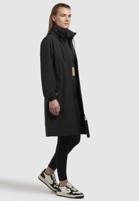 Cappotto lungo nero con colletto alto, chiusura con zip, polsini elasticizzati e tasche laterali, abbinato a leggings neri e sneakers bianco e nere.