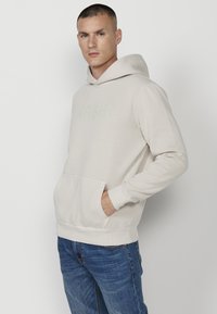 Felpa beige con cappuccio, tasca frontale e logo "KRSH" ricamato. Tessuto morbido, vestibilità rilassata, dettagli con bordo a costine alle maniche e all'orlo.