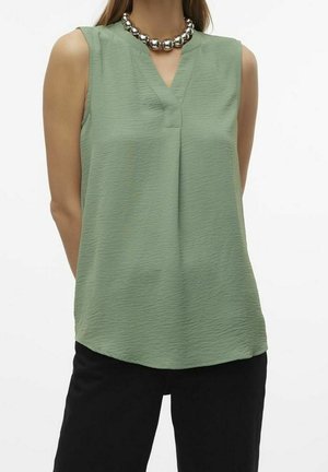 Kobieta w bezrękawowej, jasnozielonej, teksturowanej bluzce z dekoltem w szpic i srebrnym naszyjnikiem z grubych koralików, w połączeniu z czarnymi spodniami.