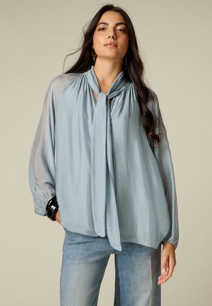 Frau mit langen dunklen Haaren, die eine lockere hellblaue Bluse mit vorderem Bindedetail und hellblaue Jeans trägt, steht vor einem schlichten Hintergrund.