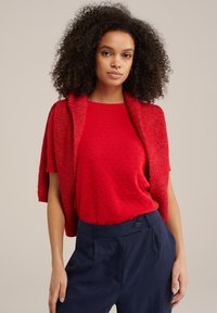 Punane lühikeste varrukatega tekstuuriga kampsun, mille peale on pandud sobiv kudumiskardigan, koos mereväe siniste kõrge vöökohaga pükste ja nööbiga detailiga.