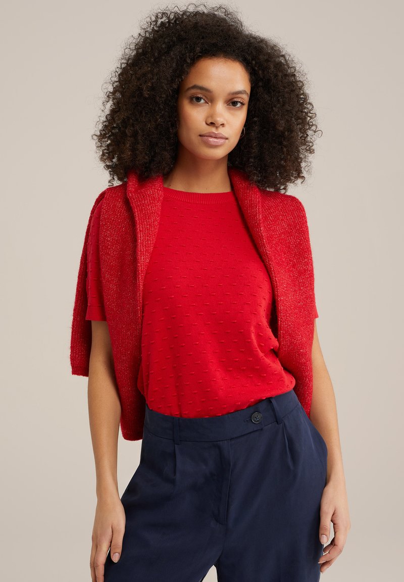 Punane lühikeste varrukatega tekstuuriga kampsun, mille peale on pandud sobiv kudumiskardigan, koos mereväe siniste kõrge vöökohaga pükste ja nööbiga detailiga.