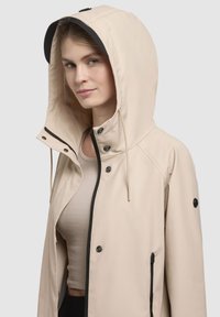 Beige waterdichte jas met een grote capuchon, drukknoopsluiting en verstelbare trekkoorden. Voorzien van een zwarte rits en zijzakken.