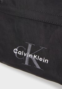 Sac en tissu noir avec logo "Calvin Klein" brodé en relief en blanc et gris. Texture lisse avec un léger éclat, design minimaliste.