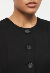 Κοντινό πλάνο του κάτω μέρους του προσώπου και του λαιμού ενός ατόμου που φοράει ένα μαύρο ένδυμα με κουμπιά με τρία ορατά μεγάλα μαύρα κουμπιά.