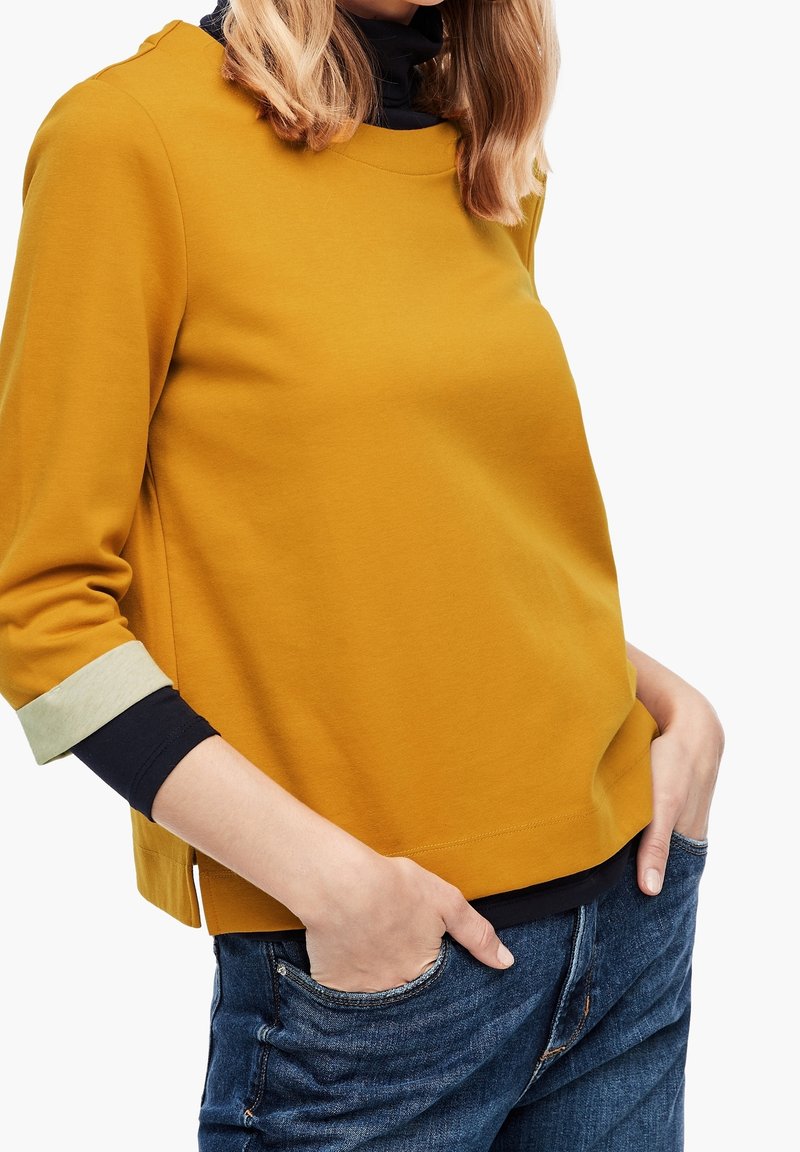 Sweat-shirt jaune moutarde à manches trois-quarts, avec des poignets vert clair contrastants et une coupe décontractée, associé à un jean foncé.