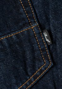 Dark blue denim fabric with orange stitching, featuring a close-up of a pocket and a black brand label partially visible.