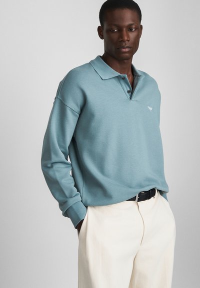 Lichtblauw poloshirt van zachte stof met een drieknoopsluiting, voorzien van geribbelde manchetten en een klein logo op de borst, gecombineerd met crèmekleurige broeken.