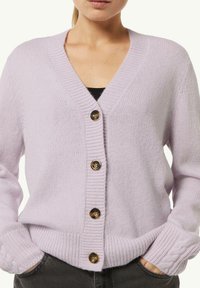 Lila Strickjacke mit V-Ausschnitt, fünf dunklen Knöpfen, gerippten Bündchen und Saum sowie einer weichen Textur. Über einem schwarzen Oberteil getragen.