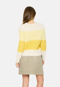 Kvinna med långt brunt hår som bär en stickad tröja i krämfärg och gult med ränder samt en beige kjol, vänd bort från kameran mot en vit bakgrund.