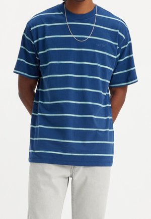 Mann trägt ein blau gestreiftes Kurzarm-T-Shirt mit hellblauen horizontalen Linien und hellen Hosen, die Hände hinter dem Rücken.