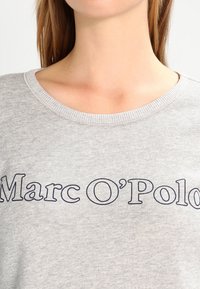 Sudadera gris hecha de una tela suave, con un escote redondo y texto azul marino "Marc O'Polo" en el pecho. Diseño clásico y casual.