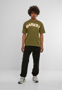 Camiseta verde oliva con "MARKET" impreso en una fuente crema contrastante, combinada con pantalones de chándal negros y zapatillas grises. Diseño y ajuste sencillos.