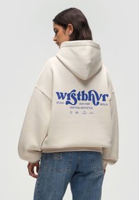 Cremfarbener Oversized-Hoodie mit Kapuze, auf dem Rücken mit blauer Grafikschrift versehen. Kombiniert mit hellblauen Jeans aus Denim, die ein gesticktes Logo zeigen.