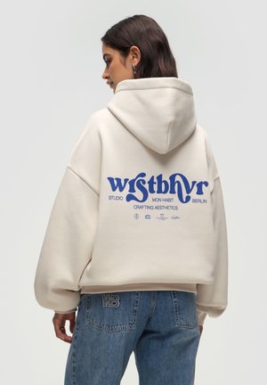 Crème oversized hoodie met een capuchon, met blauwe grafische tekst op de achterkant. Gecombineerd met lichtblauwe denim jeans met een geborduurd logo.